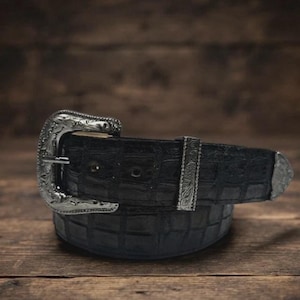 Puede incluir: Un cinturón negro con un patrón texturizado de rejilla y una hebilla de metal plateado con intrincados detalles grabados. El cinturón incluye una trabilla de metal plateado a juego y una punta de metal plateado en el extremo de la correa.