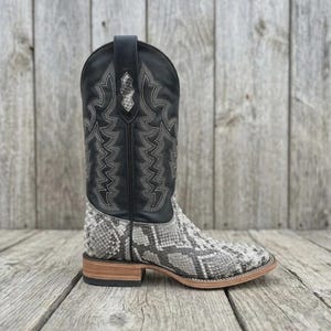 May include: A western-style boot featuring a black leather shaft with intricate grey stitching and a snakeskin-patterned foot. The boot has a square toe, a stacked wooden heel, and a pull tab at the top of the shaft with a snakeskin inlay.