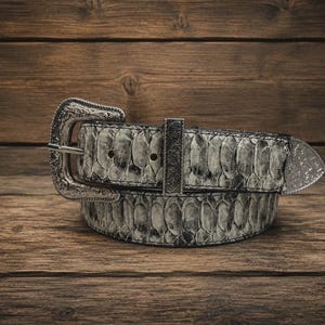May include: A belt featuring a grey and black snakeskin pattern with a silver-toned metal buckle, a matching metal belt loop, and a pointed metal tip. The buckle and hardware are decorated with intricate, embossed floral-style engravings.