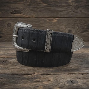 May include: A black leather belt featuring a textured surface and ornate silver-toned metal hardware. The belt includes a detailed engraved buckle, a matching silver-toned metal keeper, and a pointed silver-toned metal tip at the end of the strap.