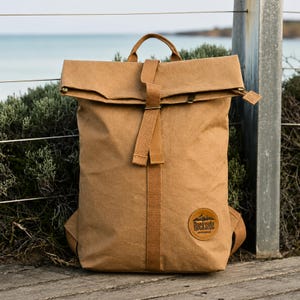 May include: A tan roll-top backpack with a textured surface, featuring a central vertical fabric strap and a circular patch on the lower right corner with the text Rockside and a mountain graphic. The bag has a top handle and shoulder straps.