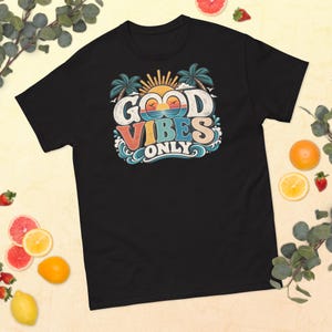 以下が含まれることがあります： GOOD VIBES ONLYというテキストがプリントされた黒の半袖Tシャツ。デザインには、黒い生地の上にオレンジ、ブルー、クリーム色のレトロなカラーパレットで描かれた、様式化された太陽、ヤシの木、海の波、鳥が描かれています。