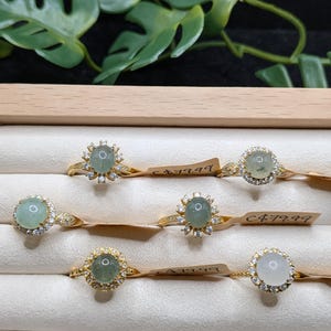 Op de afbeelding: Zes gouden ringen in een displaydoos, elk met een centrale ovale of ronde cabochon edelsteen in lichtgroene of witte tinten, omringd door een halo van kleine heldere steentjes. Elke ring heeft een label met de tekst C$79.99 eraan bevestigd.