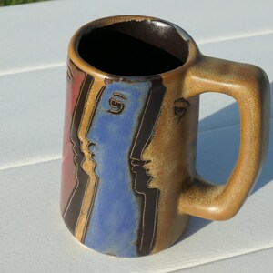 May include: A ceramic mug with a tan, brown, and blue glaze featuring a series of stylized profile faces in black line work. The mug has a thick, rectangular handle and a dark brown interior. The exterior surface displays a smooth, glossy finish.