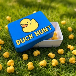 Könnte beinhalten: Eine rechteckige blaue Aufbewahrungsbox mit einer gelben Quietscheentchen-Grafik und dem auf den Deckel gedruckten Text DUCK HUNT. Die Box ist leicht geöffnet, zeigt ein weißes Inneres und ist von zahlreichen kleinen gelben Entenfiguren umgeben.