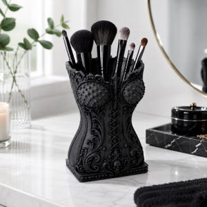 Puede incluir: Un soporte para brochas de maquillaje en negro mate con forma de corsé, adornado con volutas intrincadas, un diseño de cordones central y un pequeño detalle de calavera en la base. Contiene seis brochas con mangos negros y cerdas suaves.