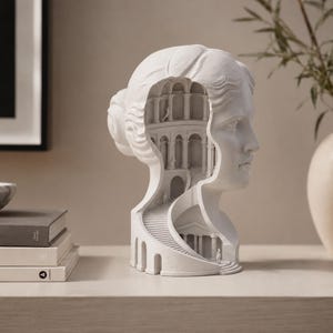 Könnte beinhalten: Eine weiße, matte Skulptur eines menschlichen Kopfes im Profil mit einem ausgehöhlten Inneren, das eine detaillierte architektonische Szene mit mehreren Ebenen, Bogensäulen und einer gewundenen Wendeltreppe zur Basis der Büste zeigt.