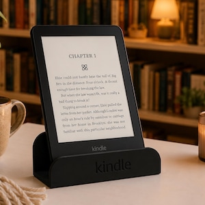 Può includere: Un lettore e-reader Kindle nero appoggiato su un supporto in plastica nera abbinato. Lo schermo mostra un testo che inizia con Chapter 1, con un paragrafo su un personaggio di nome Elsie. La parola Kindle è stampata sul dispositivo e sul supporto.