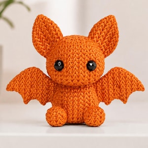 Pode incluir: Um pequeno morcego de pelúcia tricotado em laranja brilhante com uma construção de fio texturizado, duas orelhas grandes e pontiagudas, asas estendidas e dois olhos redondos de plástico preto brilhante. A criatura está sentada com pés visíveis.