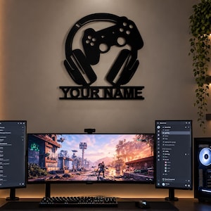 Könnte beinhalten: Ein schwarzes Wand-Schild mit der Silhouette von Gaming-Kopfhörern und einem Gamecontroller, mit einem horizontalen Balken darunter, der den Text YOUR NAME in einer fetten serifenlosen Schrift anzeigt, über einem Computer-Setup.