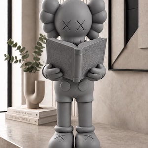 Puede incluir: Una figura KAWS de color gris mate en posición vertical, que muestra al personaje icónico con ojos en forma de X y manos sosteniendo un libro abierto. La figura tiene orejas redondeadas, pantalones cortos con dos botones y zapatos grandes.