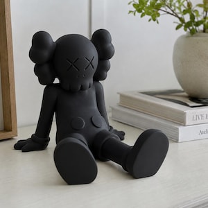 May include: A matte black vinyl collectible figure in a seated position with crossed-out eyes and bone-like ears. The character features a skull-shaped head, rounded torso buttons, and large, smooth feet, presented as a solid, monochromatic sculpture.