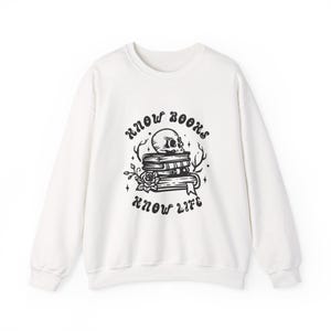 May include: A white long-sleeve sweatshirt featuring a black graphic print of a stack of books topped with a human skull, surrounded by a rose and branches. The text "KNOW BOOKS KNOW LIFE" is printed in a curved font above and below the graphic.