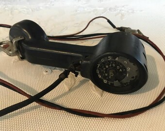 May include: A black rotary dial telephone handset with a black cord. The dial has numbers 1 through 9 and 0, and letters A through Z.