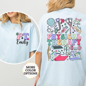 May include: A light blue t-shirt featuring a small graphic on the front left chest with the text PT Emily and a large colorful graphic on the back with the text PHYSICAL THERAPY surrounded by illustrations of exercise equipment and medical tools.