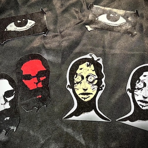 May include: A collection of six stickers featuring artistic illustrations. Two rectangular stickers show single eyes. Four die-cut stickers depict stylized human-like faces, including one with a red face, one with a yellow face, and two in black and white.