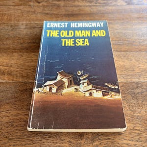 May include: A paperback book titled The Old Man and the Sea by Ernest Hemingway. The cover features a dark blue sky over a brown shoreline with small wooden huts and several small boats floating on the water. The title is in bold yellow text.