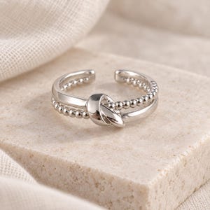 May include: A silver-toned open ring featuring a central knot design. The band consists of two parallel segments, one with a smooth polished finish and the other with a beaded texture, meeting at a central knot that connects the two sides.