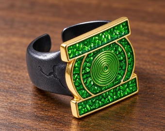 May include: A ring featuring a matte black band with a textured surface and a gold-toned rectangular emblem. The emblem displays a central green circular spiral surrounded by a vibrant, faceted green inlay within a gold frame.