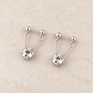 May include: A pair of silver-toned barbell earrings featuring a straight post with a ball at each end. A delicate chain connects the two ends of each barbell, suspending a small, polished, twisted knot-shaped charm in the center of the chain.