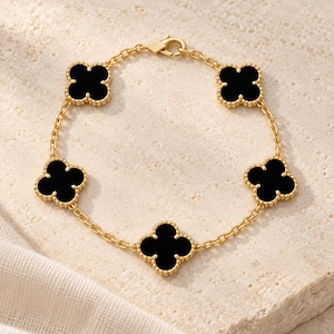 May include: A gold chain bracelet featuring five evenly spaced black clover-shaped charms with beaded edges. The bracelet is secured with a lobster claw clasp and is constructed from polished gold-tone metal links connecting each of the floral motifs.