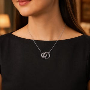 Puede incluir: Un collar de tono plateado con dos colgantes circulares entrelazados suspendidos de una cadena fina. Los anillos tienen un acabado metálico pulido y están conectados para formar un punto focal sobre una prenda de vestir de color negro liso.