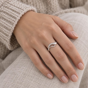 Puede incluir: Un anillo de tono plateado con forma de serpiente enroscada, que presenta un patrón de escamas texturizado a lo largo del cuerpo y una cola curva y lisa que se envuelve alrededor del dedo. El metal tiene un acabado pulido que refleja la luz.