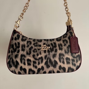 Könnte beinhalten: Eine Coach-Schultertasche mit beigefarbenem und schwarzem Leopardenmuster sowie dunkelburgunderfarbenen Lederbesätzen. Die Tasche verfügt über einen goldfarbenen Kettenriemen, ein goldenes Coach-Logo auf der Vorderseite und einen passenden Anhänger aus burgunderfarbenem Leder an der Seite.