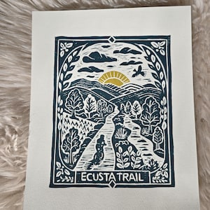 May include: A rectangular art print on off-white paper featuring a dark blue linocut illustration of the Ecusta Trail. The design shows a cyclist on a path, trees, mountains, clouds, a golden sun, and a bird, all framed by a decorative border.