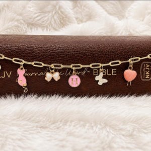 May include: A gold-toned chain bracelet featuring seven hanging charms: a white heart, a pink cat, a white bow, a pink circle with the letter H, a white butterfly, a pink heart, and a white circle with a brown cross, positioned in front of a brown book.