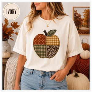 May include: An ivory short-sleeved t-shirt featuring a large apple graphic on the chest. The apple is divided into four quadrants with different patterns: floral, gingham, plaid, and leopard print, topped with a dark green leaf accent.