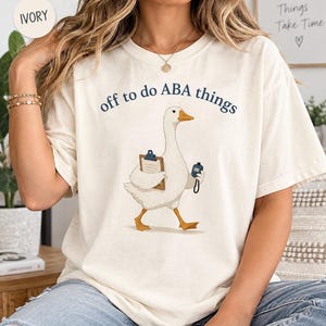 May include: An ivory t-shirt featuring a graphic of a white goose walking while holding a brown clipboard and a blue clicker. The text above the goose reads "off to do ABA things" in dark blue lettering. The shirt has a crew neckline and short sleeves.