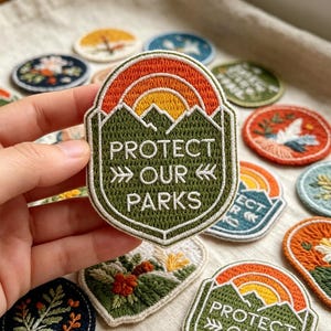 May include: An embroidered patch featuring a green mountain landscape under a yellow and orange sun, with the white text "PROTECT OUR PARKS" centered. The patch has a white border and is surrounded by various other circular and shaped embroidered patches.