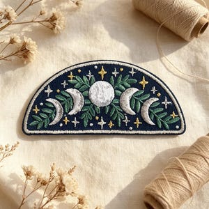 May include: A semi-circular embroidered patch featuring a dark blue background with a central white full moon, flanked by two silver crescent moons, green leafy foliage, and small yellow and white four-pointed stars throughout the design.