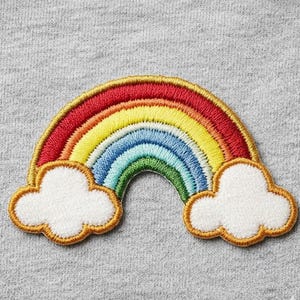 May include: An embroidered patch featuring a colorful rainbow arching over two white clouds. The rainbow consists of concentric bands in red, orange, yellow, green, and blue, all outlined with a gold-colored thread on a gray fabric background.