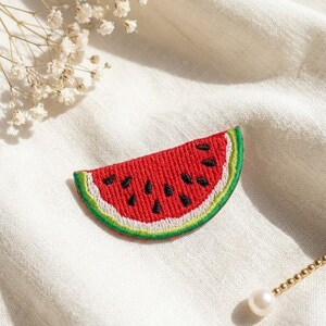 May include: A watermelon slice patch featuring red embroidery for the fruit, black stitched seeds, a thin white rind, and a green outer border. The patch is shaped like a semi-circle and displays detailed thread work throughout the design.