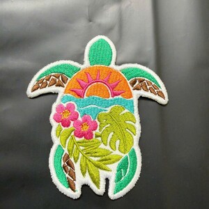 May include: An embroidered patch shaped like a sea turtle. The shell features a vibrant orange sun rising over a light blue ocean. The turtle's body is green with brown accents on the flippers, and it is decorated with two pink flowers and green leaves.