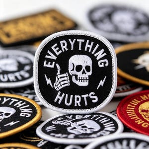 May include: A square embroidered patch with rounded corners featuring a black background, a white border, and a white skull giving a thumbs up. The text "EVERYTHING" is arched above the skull, and "HURTS" is printed below, with two small lightning bolts.