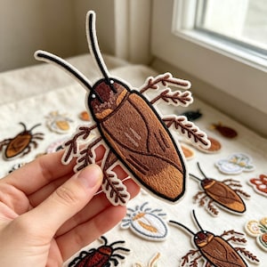 May include: An embroidered patch shaped like a brown cockroach with long black antennae and detailed legs. The insect features a dark brown head and a lighter brown body with stitched wing textures and a thick white border outline.