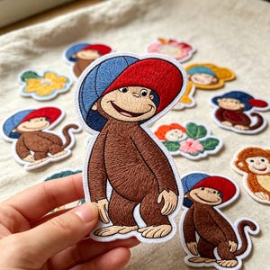 May include: An embroidered patch featuring a brown monkey wearing a blue and red baseball cap. The monkey has a light tan face, a smiling expression, and a long tail. The patch has a white border and is shown against a collection of similar patches.