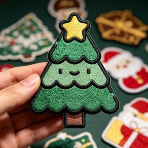 May include: An embroidered patch shaped like a stylized Christmas tree with a smiling face, featuring a bright yellow star at the top, layered green branches with black outlines, and a brown trunk, all rendered in a cartoon-like aesthetic.