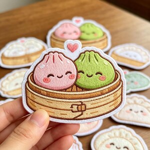 May include: An embroidered patch featuring a pink dumpling and a green dumpling sitting inside a tan bamboo steamer basket. A small pink heart is stitched above the dumplings. The patch has a white border and a cartoon face on each dumpling.