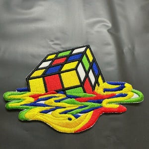 May include: An embroidered patch featuring a three-dimensional cube puzzle with red, yellow, blue, green, and white squares. The base of the cube appears to be melting into a puddle of vibrant, swirling liquid colors including yellow, blue, red, and green.