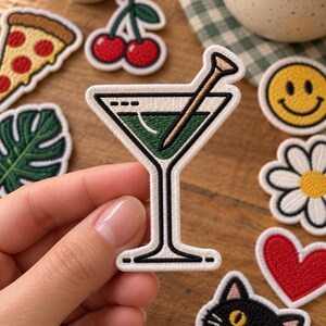 May include: An embroidered patch shaped like a martini glass filled with a green liquid and a tan cocktail pick. The patch features a white border, black outlines, and a stem base. It is held against a background of various other small embroidered patches.