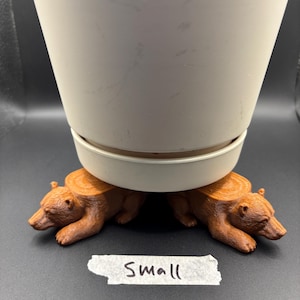 May include: Two brown bear-shaped plant pot risers supporting a white plastic flower pot. Each bear figure features a flat, circular, wood-grain textured top surface. A small piece of white tape with the handwritten word "Small" is placed in front.