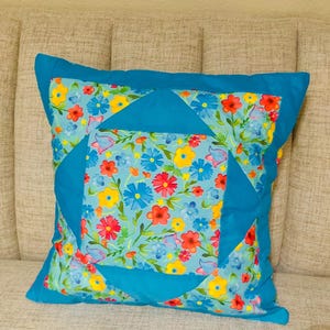 May include: A square throw pillow featuring a central panel of light blue fabric with a colorful floral print of red, yellow, and blue flowers. The design includes solid teal fabric borders and triangular accents that frame the floral center.
