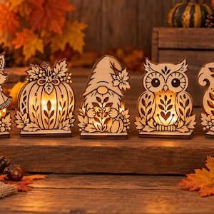 Puede incluir: Un conjunto de cinco decoraciones de madera cortadas con láser que incluyen un zorro, una calabaza, un gnomo, un búho y una ardilla. Cada pieza tiene detalles calados intrincados y está iluminada desde el interior por una luz cálida.