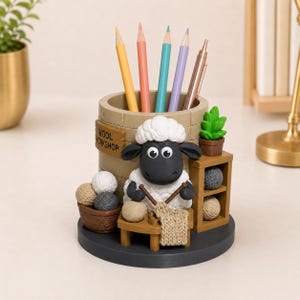 May include: A cylindrical pencil holder featuring a sculpted black and white sheep knitting with yarn. The holder has a tan brick-textured exterior with a sign reading "WOOL WORKSHOP," a small brown basket of yarn balls, and a wooden shelf with yarn.