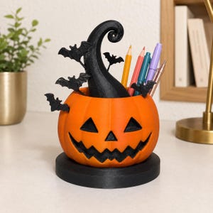 May include: A bright orange jack-o'-lantern pencil holder featuring a black swirling stem and several small black bat silhouettes attached to the sides. The pumpkin sits on a circular black base and contains several colored pencils and pens.