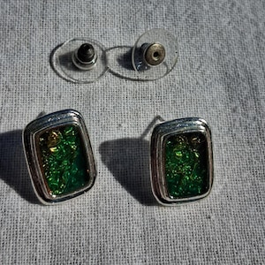May include: A pair of rectangular stud earrings featuring a metallic silver-toned frame surrounding a vibrant, textured green and gold iridescent inlay. The earrings include clear plastic disc backings for secure attachment to the post mechanism.